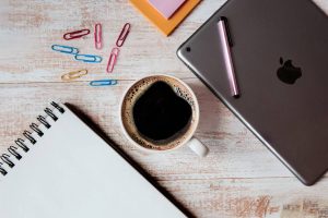 A desk with a cup of coffee, notebook, laptop, paper clips, and a pen.