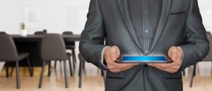 A businessman with a black suit and black shirt holds out an iPad. The businessman is only shown from the neck to the waist. Rows of black chairs are behind the businessman.