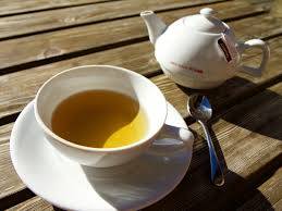 A white teacup filled with tea sits atop a white saucer. Next to the teacup are a silver spoon and white teapot.
