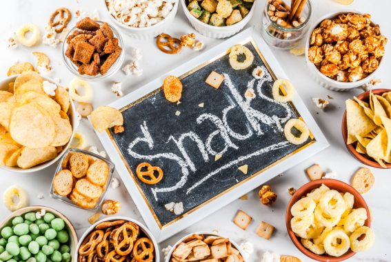 The word "snacks" written on a chalkboard with many snacks in bowls around it